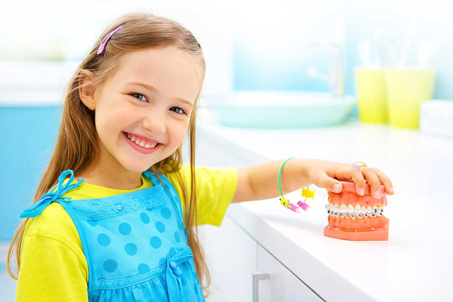 Symbolbild: Ein lächelndes Mädchen im Grundschulalter hält ein Zahnmodell mit fester Zahnspange. (Illustration)